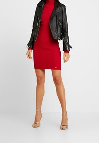 Schwarze Leder-Motorradjacke über einem figurbetonten roten Rippstrickkleid. Das Outfit wird mit silbernen, mit Riemchen versehenen High Heels kombiniert.