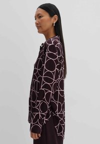 Long-sleeve shirt in dark burgundy with large pink circular patterns, featuring a relaxed silhouette and button closure at the collar.