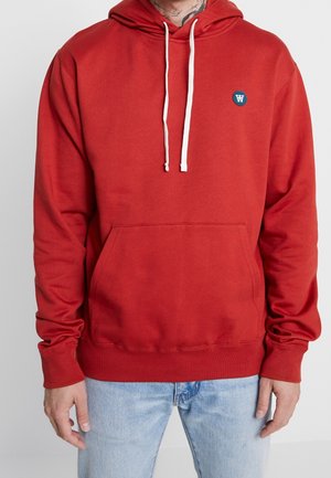 Osoba w czerwonej bluzie z kapturem i białymi ściągaczami oraz małym okrągłym logo na piersi, w połączeniu z jasnoniebieskimi dżinsami.