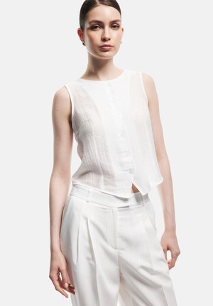 Joven mujer vestida con blusa blanca sin mangas abotonada y pantalones plisados blancos de talle alto, de pie con postura relajada sobre fondo blanco.