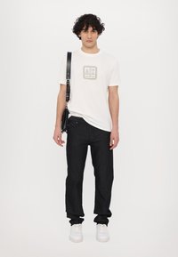 White cotton t-shirt with a logo and black jeans. The model holds a black crossbody bag and wears white sneakers. Plain white background.
