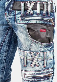 Jean en denim avec un délavage bleu, présentant des accents en cuir noir, des surpiqûres rouges, plusieurs poches et une fermeture éclair. Étiquette de marque visible.