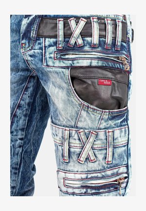 Denim-Jeans mit einer ausgewaschenen blauen Waschung, ausgestattet mit schwarzen Lederakzenten, roten Nähten, mehreren Taschen und einem Reißverschluss. Markenlabel sichtbar.