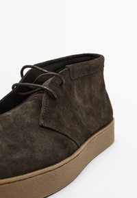 Bottines en suede marron avec une finition texturée, présentant un bout arrondi, une semelle en caoutchouc beige contrastante, et des lacets noirs.