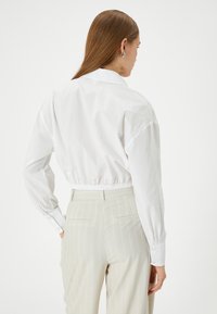 Camicetta bianca corta con maniche lunghe, vita elasticizzata e colletto a punta. Indossata con pantaloni a righe beige chiaro, che mostrano una texture liscia.