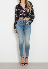 Žena oblečená v zesvětlených modrých skinny džínech s vysokým pasem, černé květinové a tečkované zkrácené košili s uzlem, černých lodičkách na kotníky a perlovém náhrdelníku.