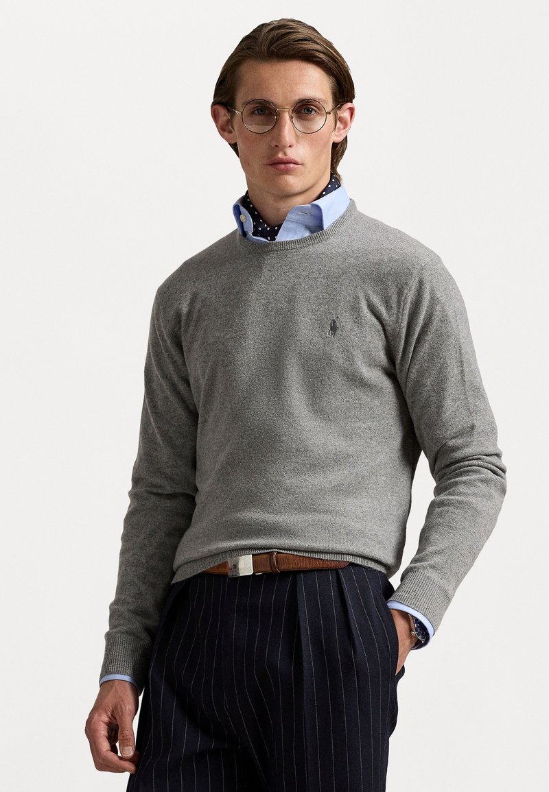 Pull en tricot gris avec un col rond et un petit logo sur la poitrine, porté par-dessus une chemise à col bleu clair avec des pois navy.