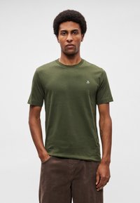 Πράσινο λαδί βαμβακερό T-shirt με κοντά μανίκια, στρογγυλή λαιμόκοψη και μικρό λευκό λογότυπο στο αριστερό στήθος. Υφή υφάσματος και μοντέρνα εφαρμογή.