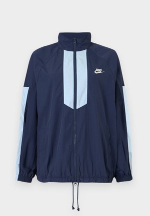 Marineblå vindjakke med lyseblå detaljer, høj krave, fuld frontlynlås og elastiske manchetter. Nike-logo på brystet.