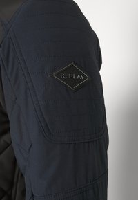 Veste matelassée bleu marine en tissu texturé, avec un patch noir en forme de losange portant l'inscription « REPLAY » sur l'épaule gauche.