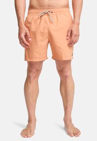 Άνδρας φοράει πορτοκαλί μαγιό με λευκό και μαύρο κορδόνι, όρθιος ξυπόλητος μπροστά σε απλό φόντο.