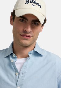 Camisa de botones azul claro con una camiseta blanca debajo y una gorra de béisbol crema con bordado en azul marino. Texturas de tela suaves en todo.