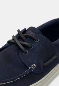 Μπλε ναυτικό δερμάτινο παπούτσι για σκάφος με καφέ κορδόνια, που διαθέτει μεταλλικά ρουθούνια και λευκή ραφή κατά μήκος της σόλας. Λεία υφή και κανονικό, καθημερινό σχέδιο.