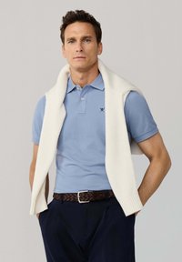 Mężczyzna ubrany w jasnoniebieską koszulkę polo, biały sweter zarzucony na ramiona, ciemne spodnie oraz brązowy pleciony pasek, stojący z rękami w kieszeniach.