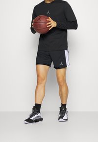 Zwarte longsleeve shirt met een logo, zwarte shorts met zijaccenten, en zwart/witte basketbalschoenen. Houdt een basketbal vast.