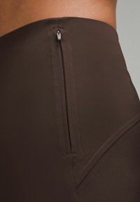 Kahvinsävyiset korkeavyötäröiset legginsit joustavasta kankaasta, joissa on sivulla vetoketjullinen tasku ja sileä, matta pinta, jossa on minimaalinen sauman määrä.