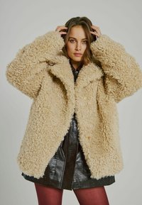 Beige kabát z umělé kožešiny s měkkým, texturovaným povrchem, širokými rukávy a uvolněným střihem, s dvoupřezkovým zapínáním a límcem.