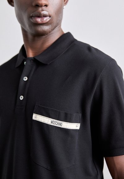 Svart polo skjorta i texturerat tyg, med en ficka med en vit remsa märkt "Moschino" och knappstängning framtill.