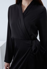 Robe noire portefeuille en tissu lisse, dotée de manches longues et d'un détail de lien sur le côté. Le design comprend un décolleté en V profond et une silhouette fluide.