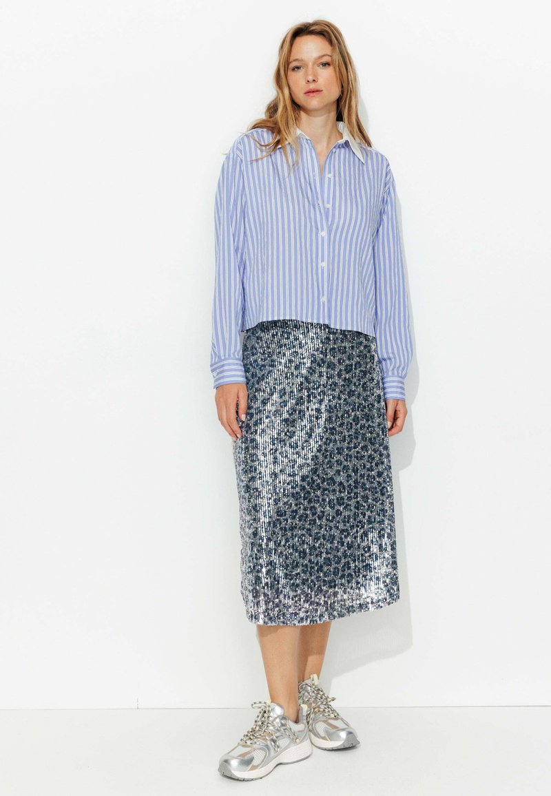 Chemise à rayures bleues et blanches à boutons, jupe midi en sequins argentés avec un motif floral, assortie à des baskets argentées. Accessoires minimalistes.