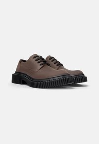 Zapatos marrones con cordones y una suela de goma negra gruesa, colocados en un ángulo para mostrar la parte delantera y lateral. El fondo es blanco liso.