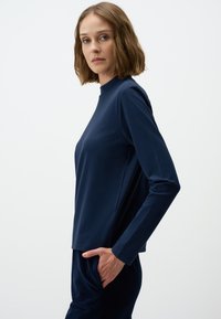 Marineblauw langemouwen bovenstuk met een hoge halslijn, gemaakt van zacht materiaal. Heeft een relaxte pasvorm en een subtiele zijnaaddetail. Gestyleerd met donkere broek.
