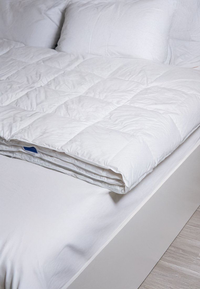 Weißer Quilt, gefaltet auf einem Bett, mit einem gesteppten Muster. Weicher Stoff mit glatter Textur. Zwei weiße Kissen im Hintergrund.