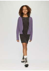 Lila Strickjacke über einem dunkelgrauen gerippten Kleid, kombiniert mit weißen Knöchelsocken und beigen Schnürstiefeln, die vor einem neutralen Hintergrund gehen.