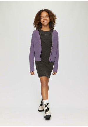 Lila Strickjacke über einem dunkelgrauen gerippten Kleid, kombiniert mit weißen Knöchelsocken und beigen Schnürstiefeln, die vor einem neutralen Hintergrund gehen.