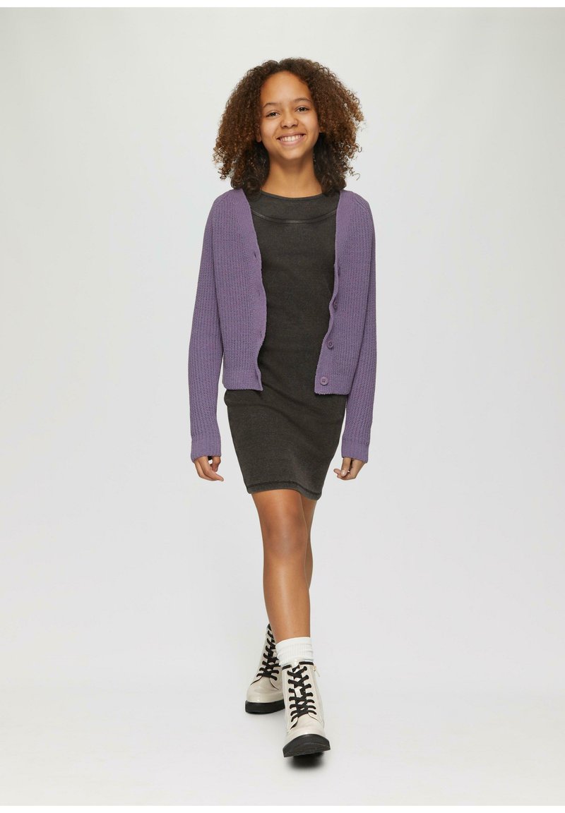 Lila Strickjacke über einem dunkelgrauen gerippten Kleid, kombiniert mit weißen Knöchelsocken und beigen Schnürstiefeln, die vor einem neutralen Hintergrund gehen.