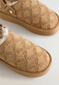 Beige Wildlederschlupfschuhe mit geprägten Blumenmustern, einem weichen Innenfutter und einer strukturierten Gummisohle mit erhöhtem Rand.