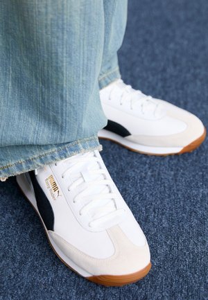 Zapatillas White Puma Easy Rider con rayas laterales negras, usadas con vaqueros azul claro sobre una alfombra azul.