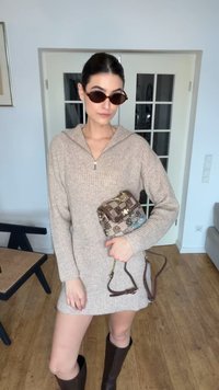 Frau trägt beiges Strickkleid und dunkle Stiefel, hält gemusterten braunen Handtasche und steht drinnen in der Nähe von weißen Türen und Holzmöbeln.