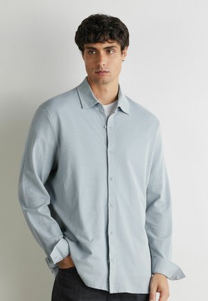 Homme aux cheveux bouclés et foncés portant une chemise bleu clair à boutons par-dessus un t-shirt blanc, debout avec une main dans la poche devant un mur uni.