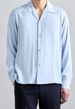 Man draagt een losse lichtblauwe, lange mouw, knoopsluitende overhemd met spreidkraag en een donkere broek, staand met één hand in zijn zak.