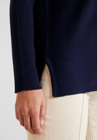 Granatowy sweter z prążkowanej dzianiny z bocznym rozcięciem na dole, w zestawie z jasnymi, teksturowanymi spodniami.