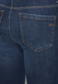 Jeans de denim azul escuro com um bolso traseiro com um pequeno logótipo amarelo "IVY" e costura em contraste. O tecido parece suave e de peso médio.