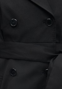 Κοντινό πλάνο ενός μαύρου παλτού διπλής σειράς με τέσσερα κουμπιά και ζώνη από ύφασμα που δένεται στη μέση.