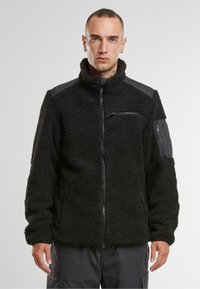 Giacca in pile nera con colletto alto, di fronte con zip e superficie testurizzata. Presenta tasche laterali e dettagli sulle spalle in un materiale liscio.