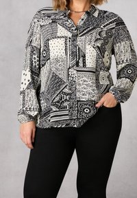 Blusa a maniche lunghe bicolore bianco e nero con colletto, caratterizzata da vari disegni geometrici e una vestibilità comoda. Pantaloni neri indossati sotto.