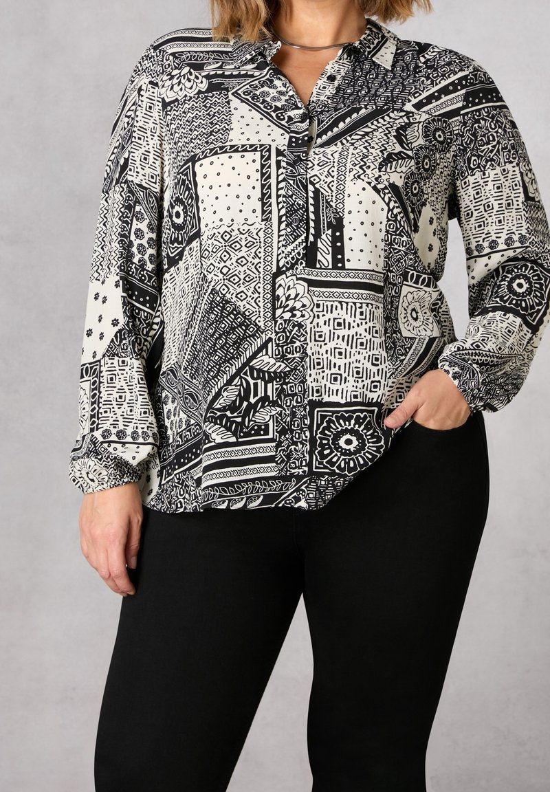 Blusa a maniche lunghe bicolore bianco e nero con colletto, caratterizzata da vari disegni geometrici e una vestibilità comoda. Pantaloni neri indossati sotto.