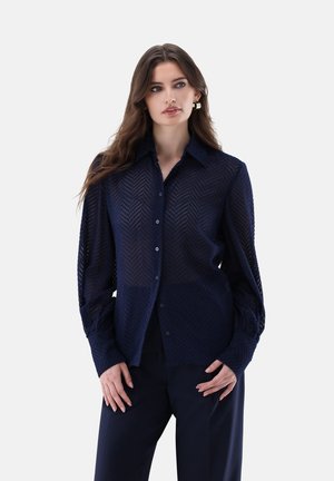 Donna con i capelli lunghi che indossa una camicetta blu scuro testurizzata con bottoni e pantaloni blu scuro abbinati, che guarda leggermente di lato.