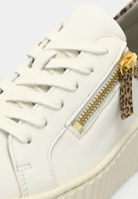 Valkoinen nahkainen lenkkari, jossa on kultainen vetoketjutekijä ja leopardikuvioinen yksityiskohta. Varustettu valkoisilla kengännauhoilla ja teksturoidulla pohjalla. Lähikuva.