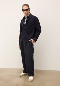 Homme portant des lunettes de soleil noires avec une veste de chemise bleu marine, une chemise à motifs gris clair, un pantalon sombre ample et des baskets blanches, se tenant devant un mur uni.