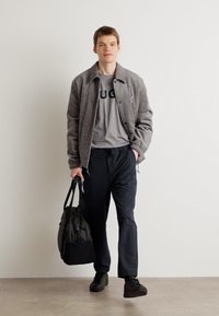 Grijze bomberjack over een grijs T-shirt met "HUGO" in het zwart, gecombineerd met zwarte broek en sneakers, met een grote zwarte tote bag.