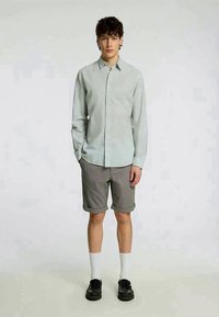 Φανελάκι σε ανοιχτό πράσινο χρώμα με μακριά μανίκια, κουμπωμένο, με γιακά; γκρι ριγέ σορτσάκι; λευκές κάλτσες μέχρι το γόνατο; μαύρα παπούτσια; απλή, casual εμφάνιση.