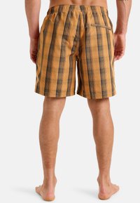 Bañador de hombre hecho de tela ligera en color naranja con un patrón de cuadros negro. Presenta una cintura elástica y un bolsillo trasero.