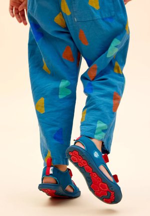 Blaue Sandalen mit roten Akzenten und strukturierten Sohlen, getragen mit blauen Hosen, die bunte herzförmige Muster in Gelb, Grün und Orange aufweisen.