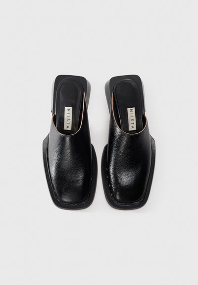 Mules en cuir noir à enfiler avec un bout carré et une semelle intérieure marquée, reposant sur une semelle épaisse et rectangulaire sur fond blanc.