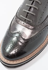 Chaussure brogue en cuir noir brillant avec des perforations détaillées, des lacets gris, une tige métallique texturée et une semelle superposée avec des coutures blanches.